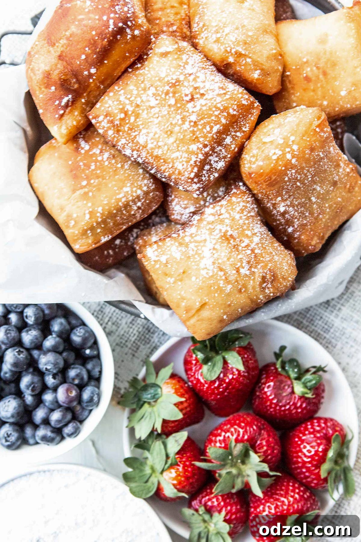 Pillowy Soft Beignet Delights 2 A bowl filled with golden-brown beignets generously dusted with powdered sugar, accompanied by small bowls of fresh strawberries and blueberries.