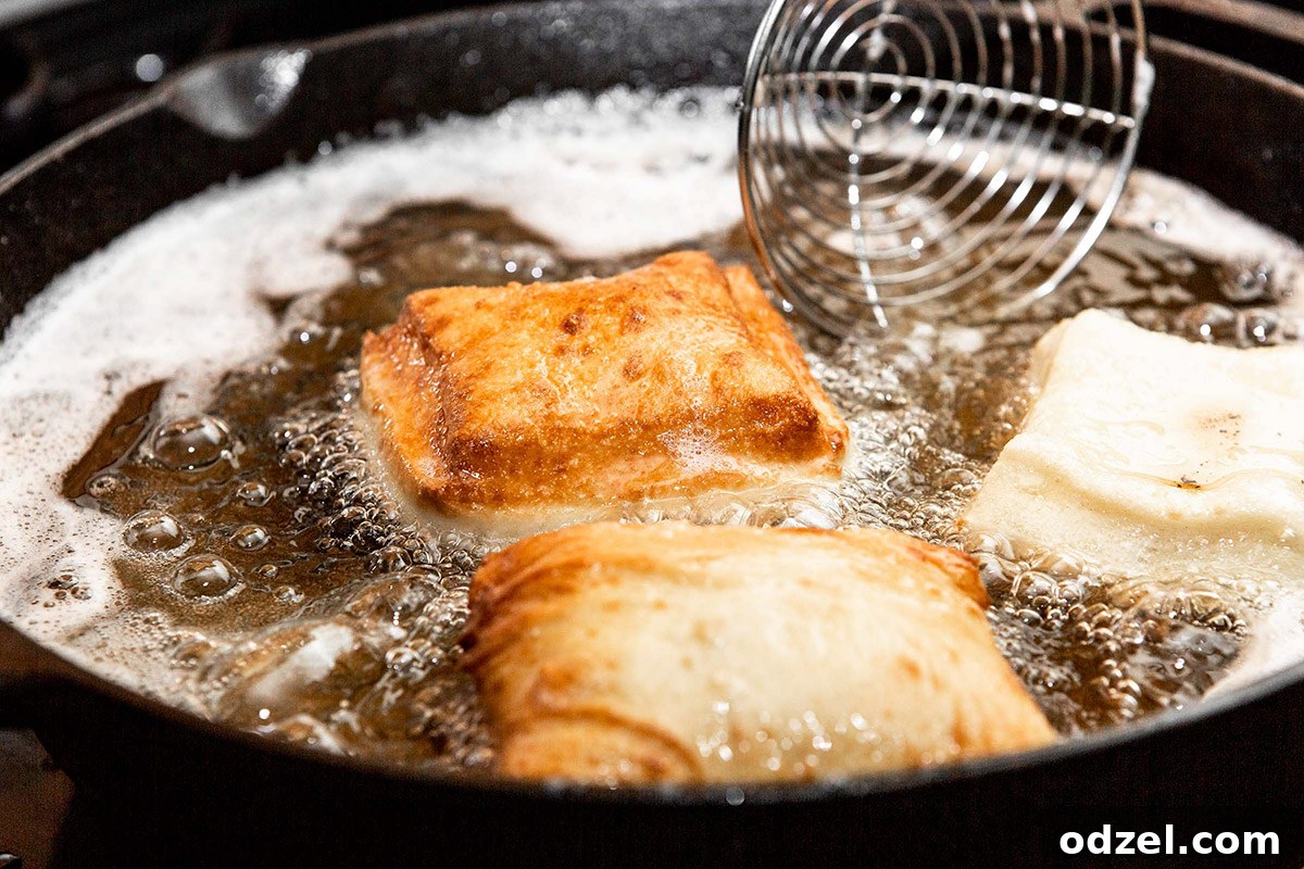 Pillowy Soft Beignet Delights 4 Three golden-brown beignets sizzling and puffing up in hot oil within a large skillet, with a round mesh skimmer nearby for removal.