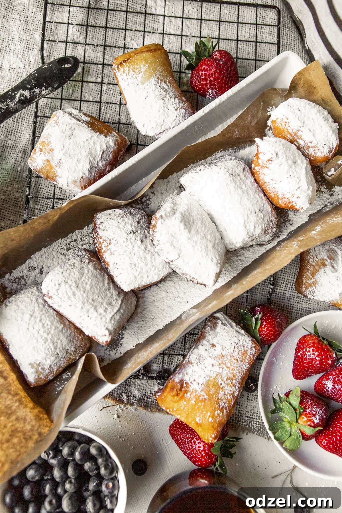 Pillowy Soft Beignet Delights 5 A long brown paper bag filled with hot, freshly fried beignets. More beignets are scattered around the bag, alongside vibrant fresh strawberries and blueberries, creating an inviting display.