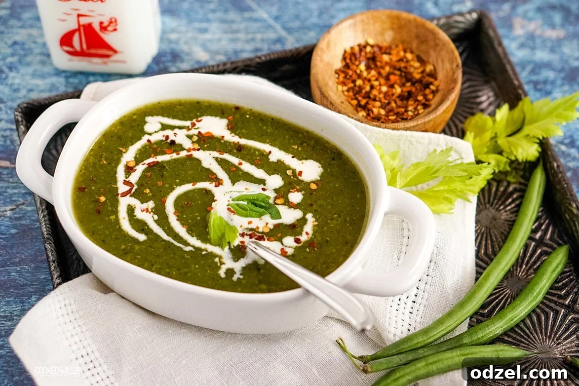 A close-up of green detox soup garnished with red crushed pepper, with fresh green beans and herbs on the side