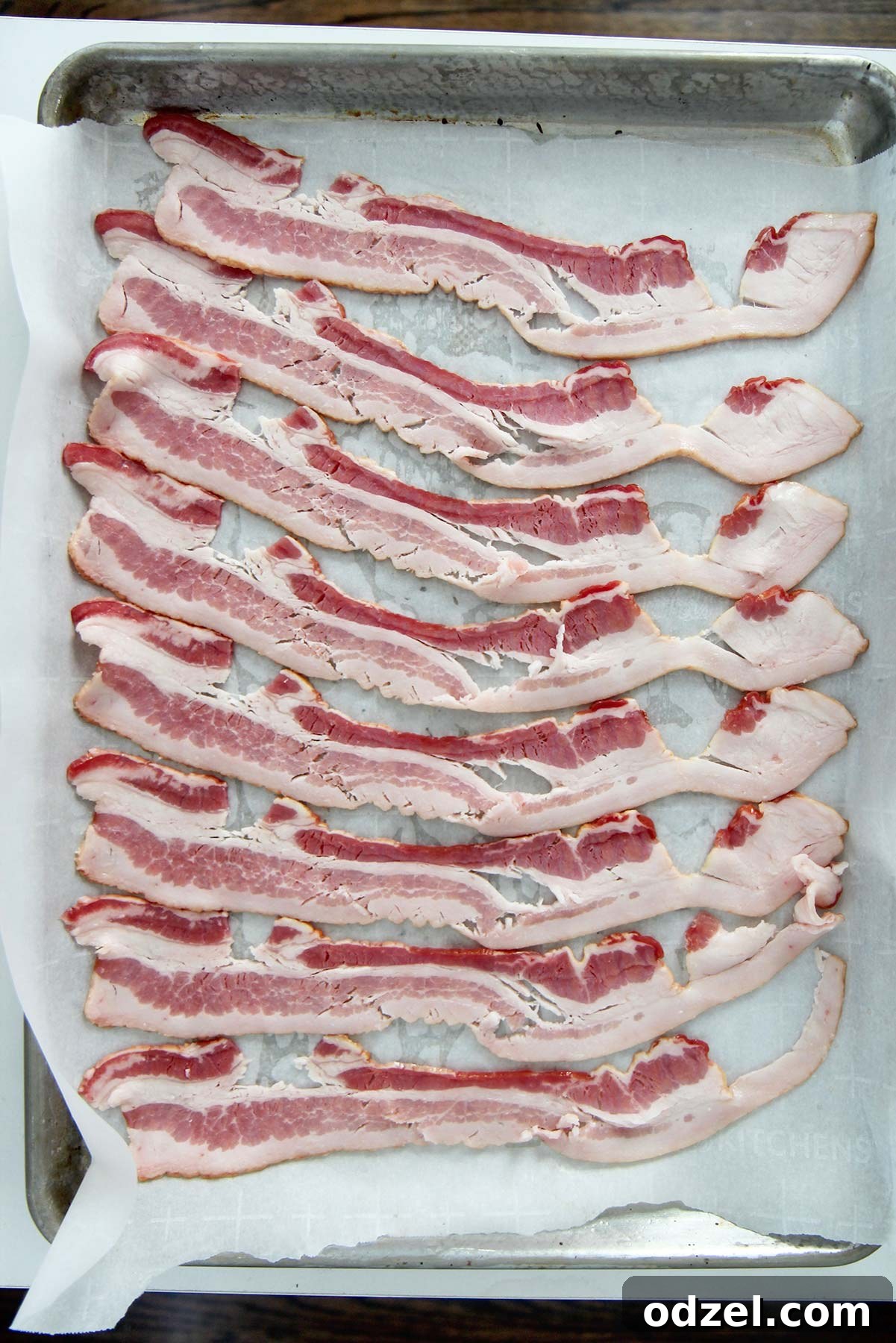 Perfectly Crispy Oven Bacon 3 Eight slices of raw bacon neatly arranged side-by-side on a baking sheet lined with parchment paper, ready for the oven.