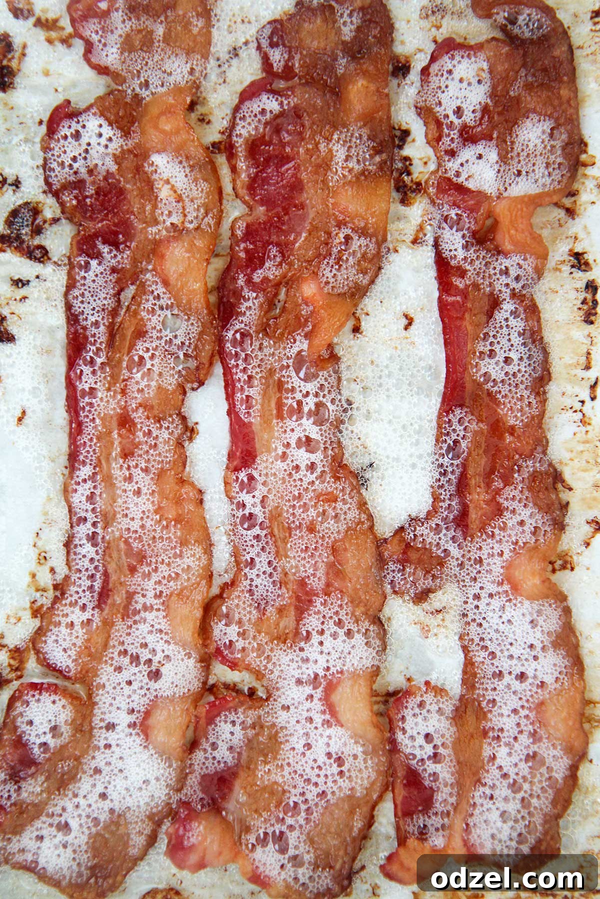 Perfectly Crispy Oven Bacon 4 Three slices of perfectly cooked, crispy bacon resting on a baking sheet, showcasing their golden-brown color and inviting texture.