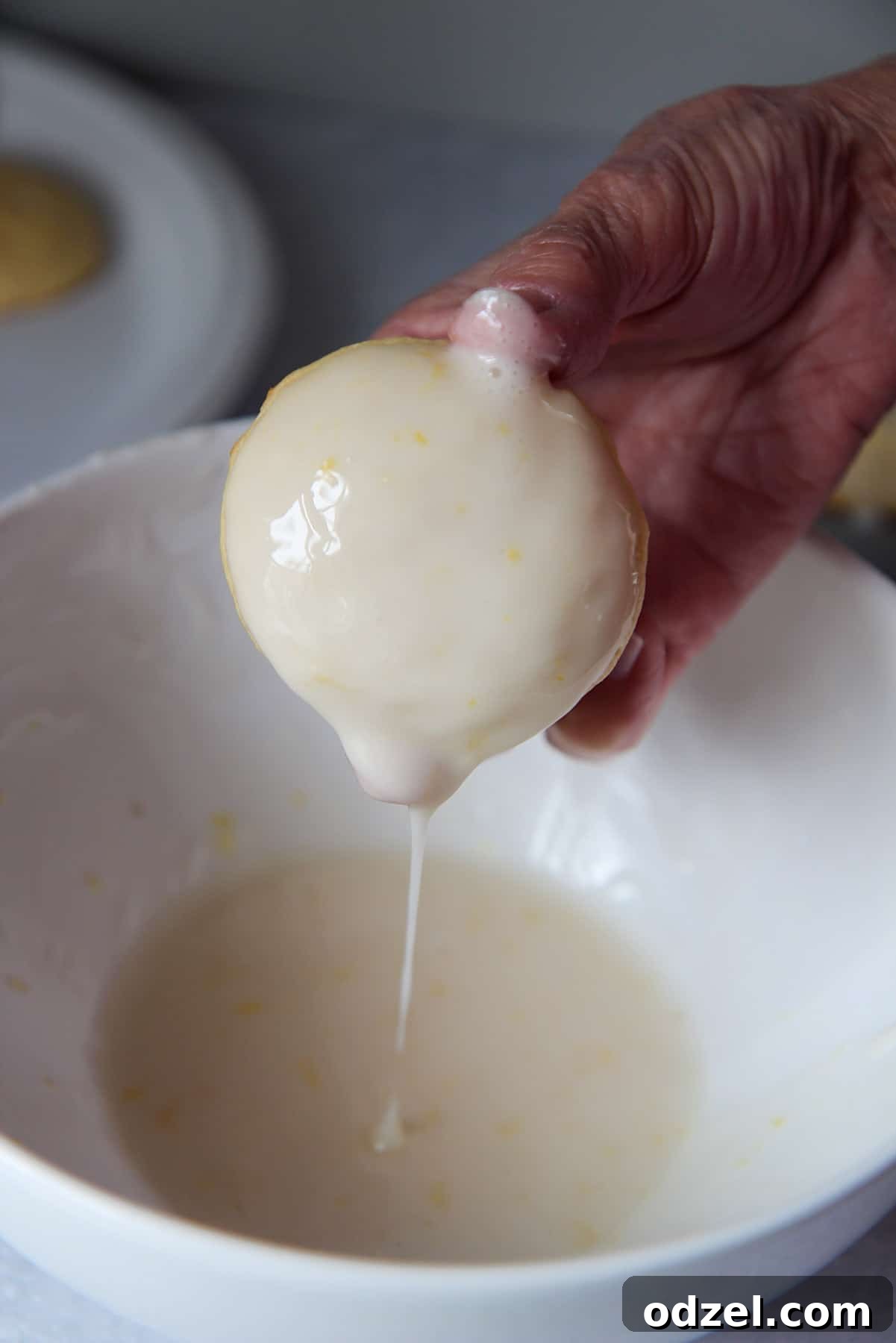 Sunshine-Kissed Lemon Ricotta Bites 5 A hand dipping a baked cookie into a white bowl filled with lemon glaze, illustrating the glazing process.