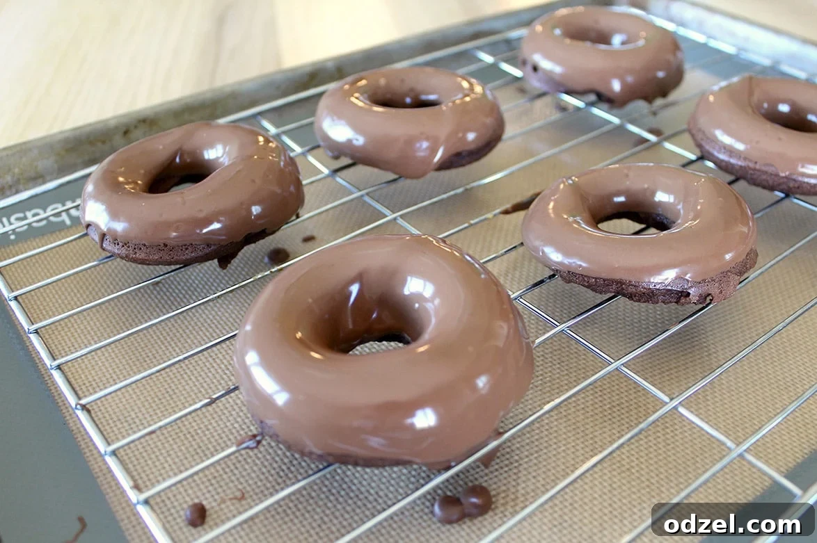 Six glossy chocolate-glazed donuts arranged neatly on a silver wire cooling rack, glistening under the light, with drops of chocolate glaze subtly catching the eye.