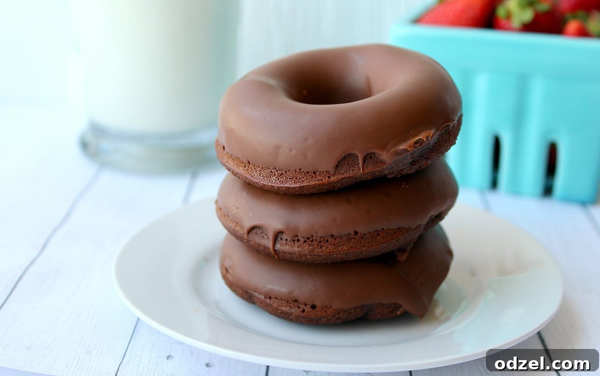 A stack of three richly glazed chocolate donuts on a white ceramic plate, artfully arranged with a glass of milk and fresh strawberries in the soft-focus background, creating an inviting dessert scene.