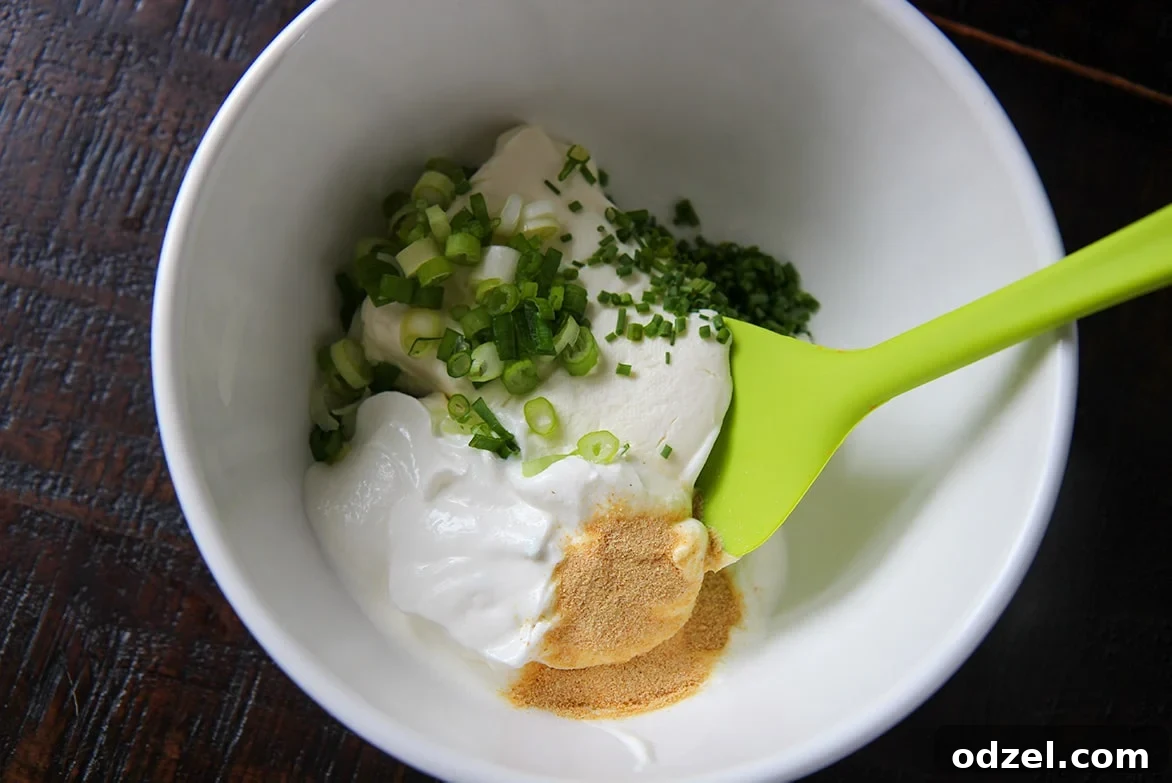 A white bowl containing softened cream cheese, sour cream, granulated garlic, fresh chives, and chopped scallions, with a green spatula ready for mixing the creamy spread.