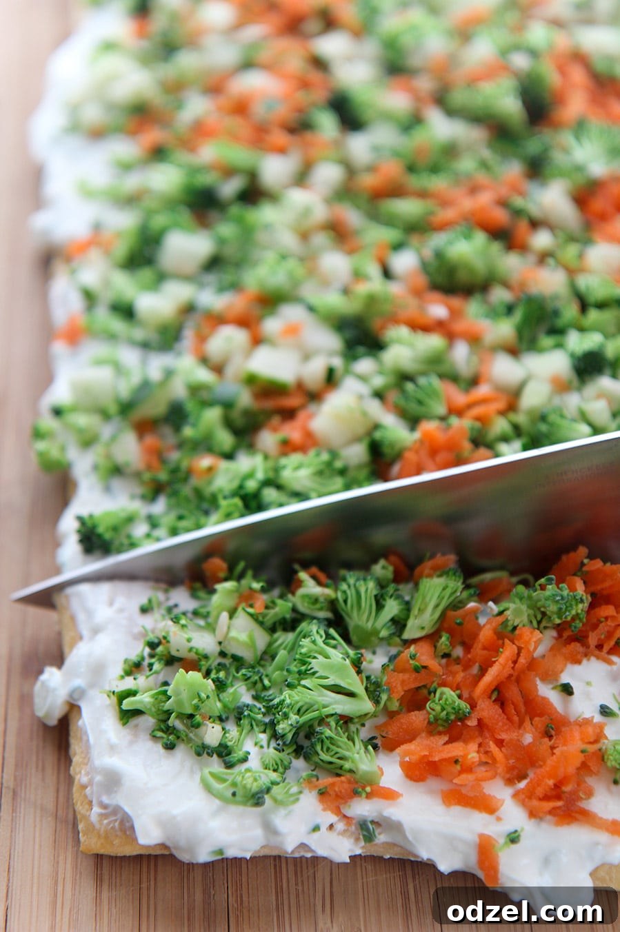 A sharp kitchen knife carefully slicing into a fully assembled pan of vibrant veggie bars, showing the distinct layers of flaky crust, creamy spread, and fresh vegetables.