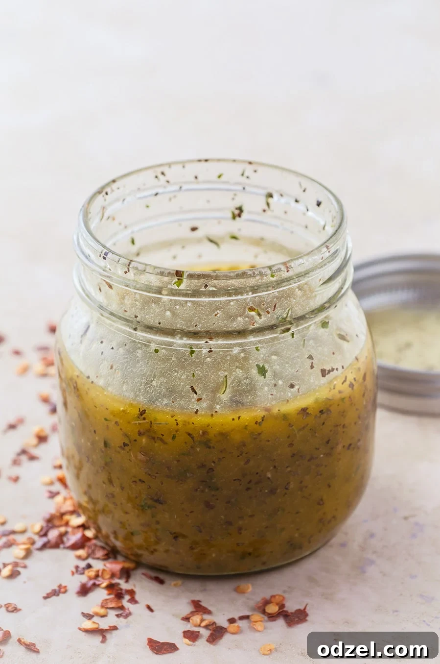 A small mason jar filled with homemade Italian dressing, with red crushed pepper and a lid on the side.