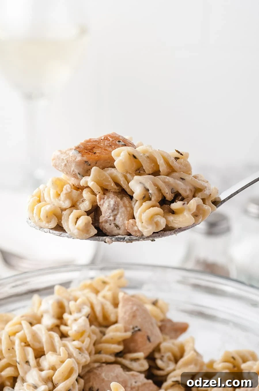 A spoon lifting a generous portion of creamy chicken and fusilli pasta from a white bowl, showing its rich texture and delicious appeal.