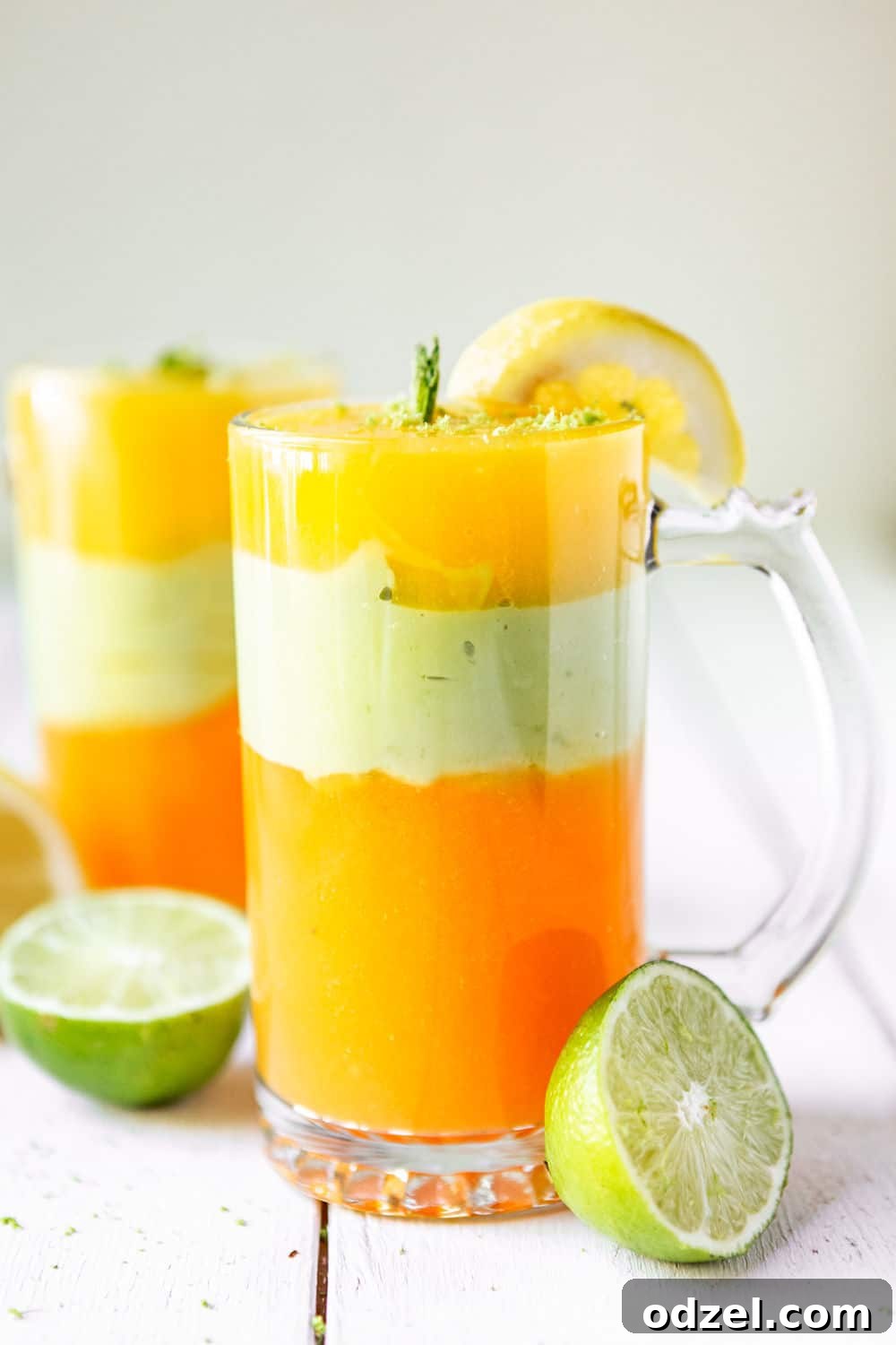 Layered Ethiopian Spris 2 Two tall glasses filled with vibrant, layered Ethiopian Spris, garnished with lime wedges and mint, on a rustic wooden surface.