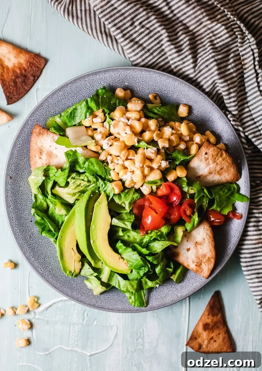 A beautifully arranged blue bowl featuring crisp lettuce, golden corn kernels, sliced avocado, and a side of crunchy tortilla chips, all resting on a stylish black and white striped towel. This image perfectly showcases the fresh and inviting Mexican Corn Salad.