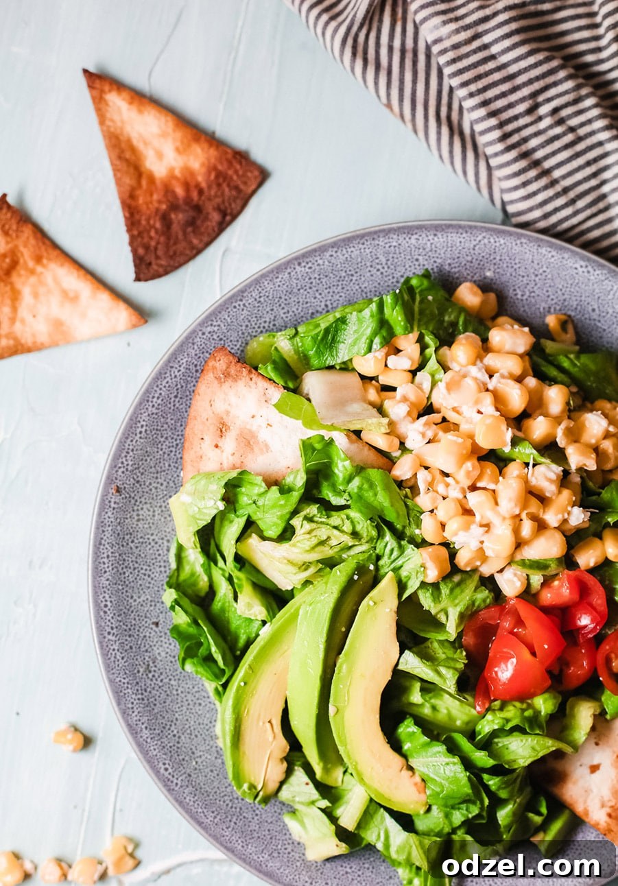 A close-up shot of a vibrant Mexican Corn Salad in a blue bowl, featuring colorful layers of lettuce, creamy avocado slices, juicy tomatoes, and a generous topping of the signature corn mixture, with tortilla chips visible on the side.