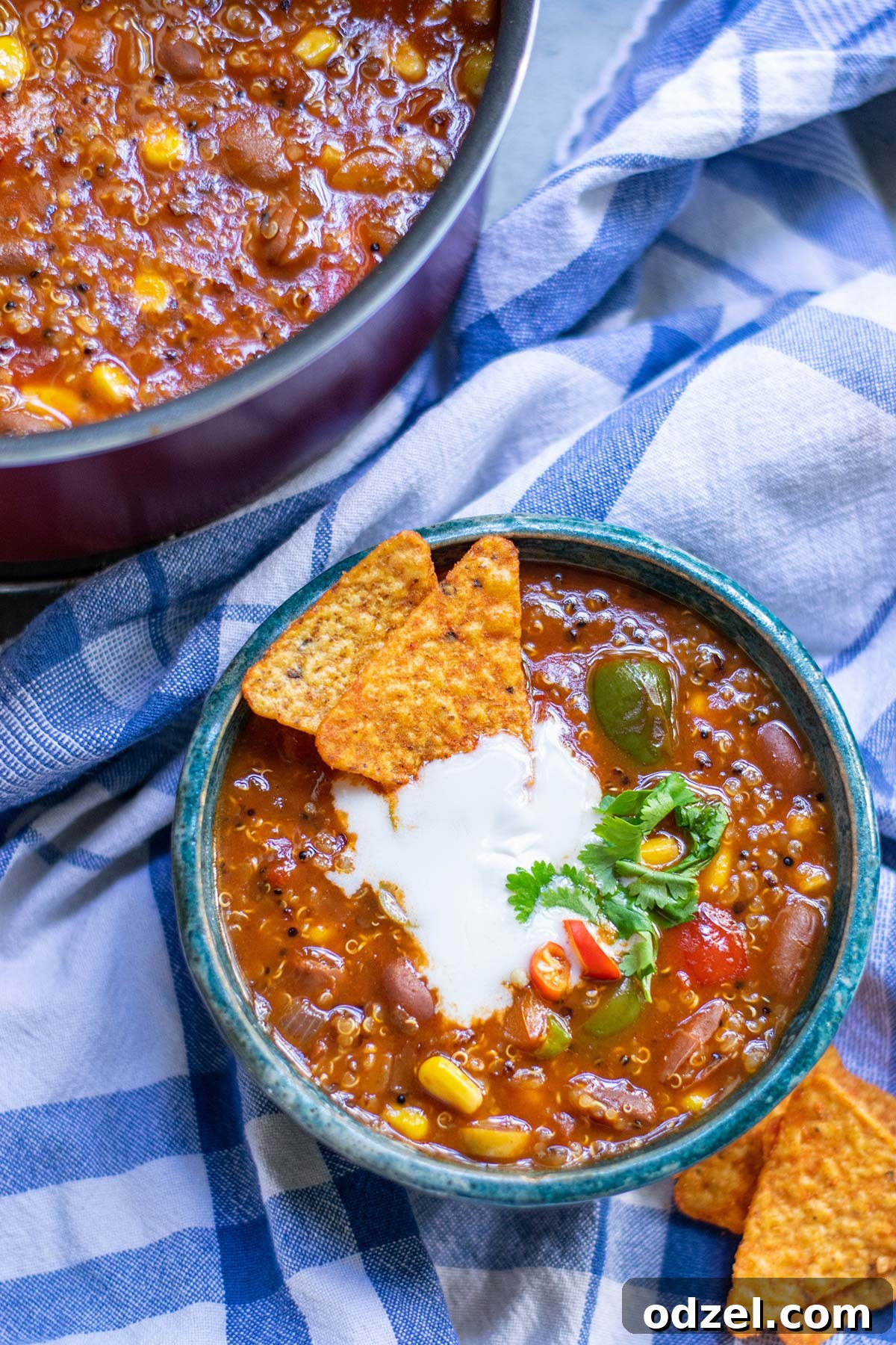 Superfood Quinoa Chili 2 A small blue bowl filled with vibrant quinoa chili, garnished with tortilla chips both on the side and within the bowl. In the background, a large pot holds more chili, all resting on a blue towel.