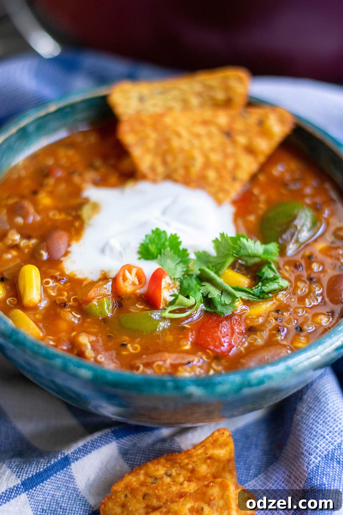 Superfood Quinoa Chili 3 A bowl filled with rich quinoa chili, elegantly topped with a dollop of white cream, fresh cilantro, and a scattering of tortilla chips, all presented on a soft blue towel.