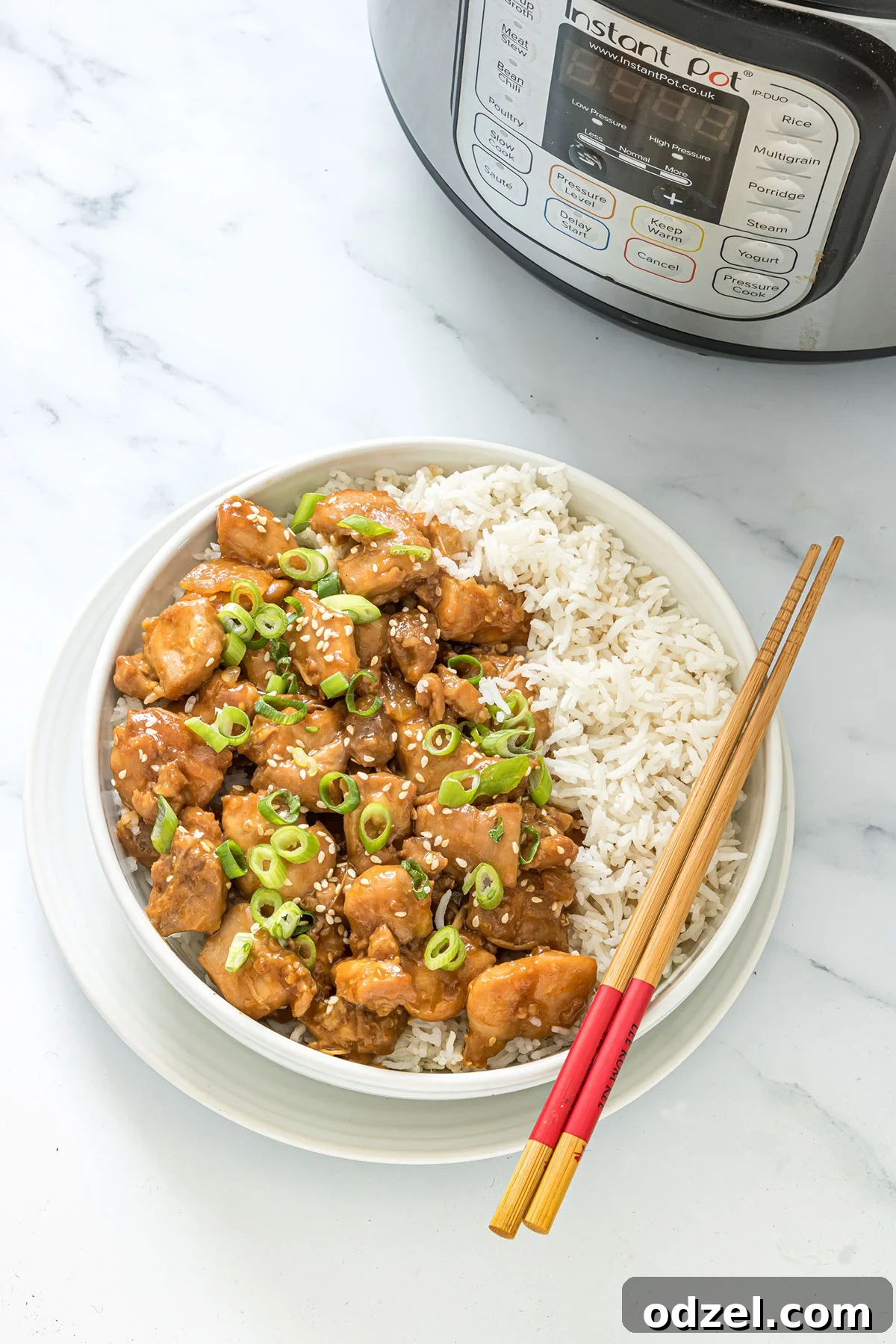 A bowl of Instant Pot sesame chicken served with white rice, garnished with green onions and sesame seeds. Chopsticks rest on the bowl, with an Instant Pot blurred in the background.
