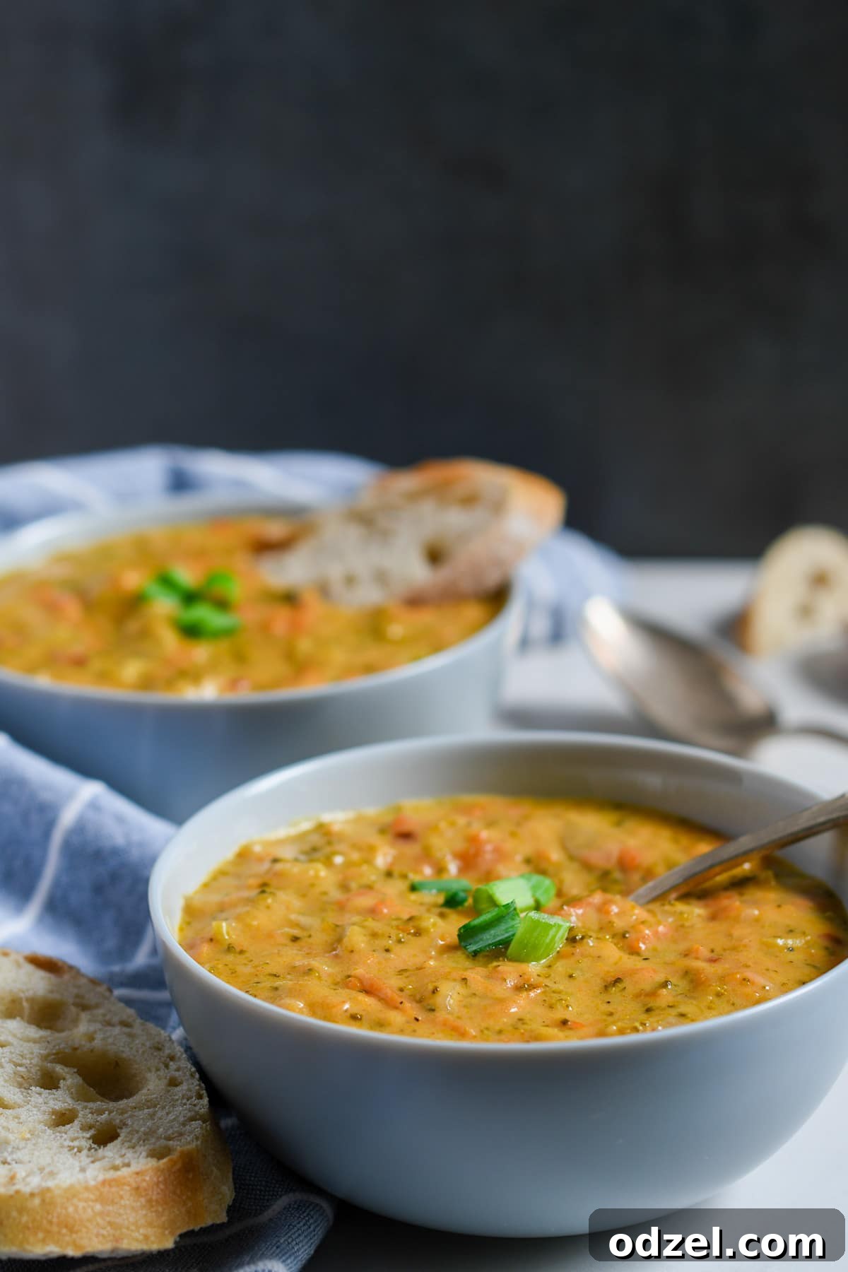 Instant Pot Creamy Broccoli Cheddar Bliss 4 Two pristine white bowls brim with creamy broccoli cheddar soup, garnished elegantly with chopped scallions. Slices of rustic bread are artfully arranged alongside the bowls, with a blue and white striped towel neatly folded in the background. The scene is set against a dark, inviting backdrop, highlighting the warmth and richness of the meal.