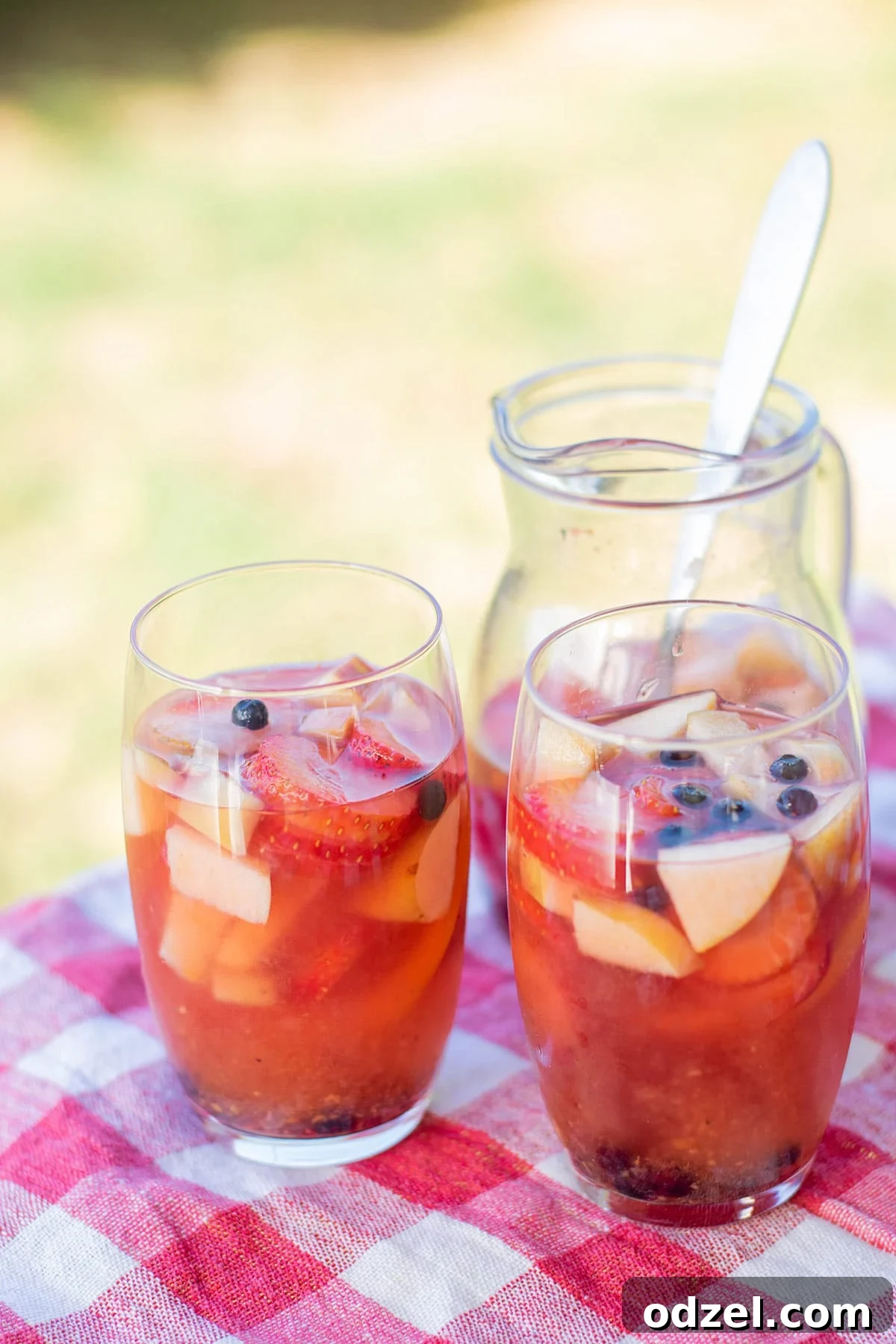 Two glasses filled with bright yellow mango sangria and fresh fruit on top, resting on a red and white checkered tablecloth. A pitcher of sangria is blurred in the background, suggesting a lively outdoor setting.