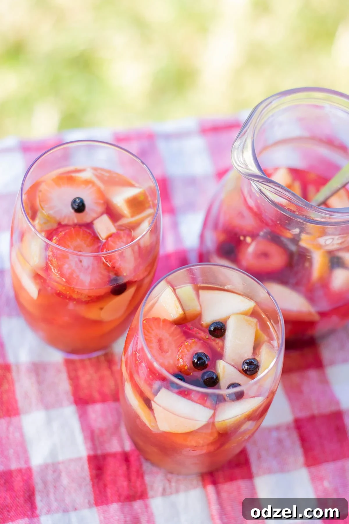 Two glasses of vibrant mango sangria, brimming with fresh fruit, are placed on a rustic picnic table outdoors. A large pitcher of sangria stands ready in the soft focus background, evoking a leisurely summer gathering.