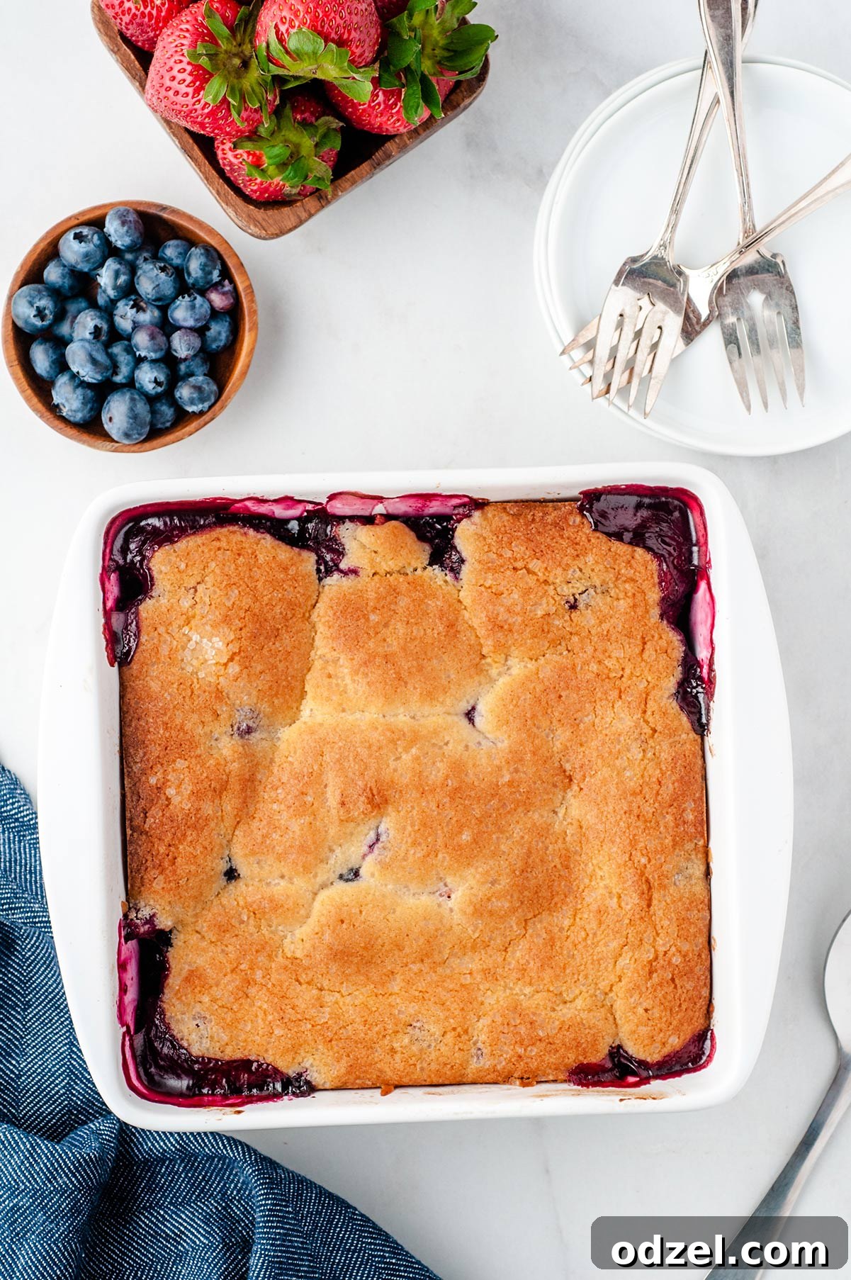 A beautifully baked berry cobbler in a white baking dish, garnished with fresh strawberries and blueberries on the side. Forks and a small white plate are thoughtfully arranged next to it, inviting a delicious spoonful.