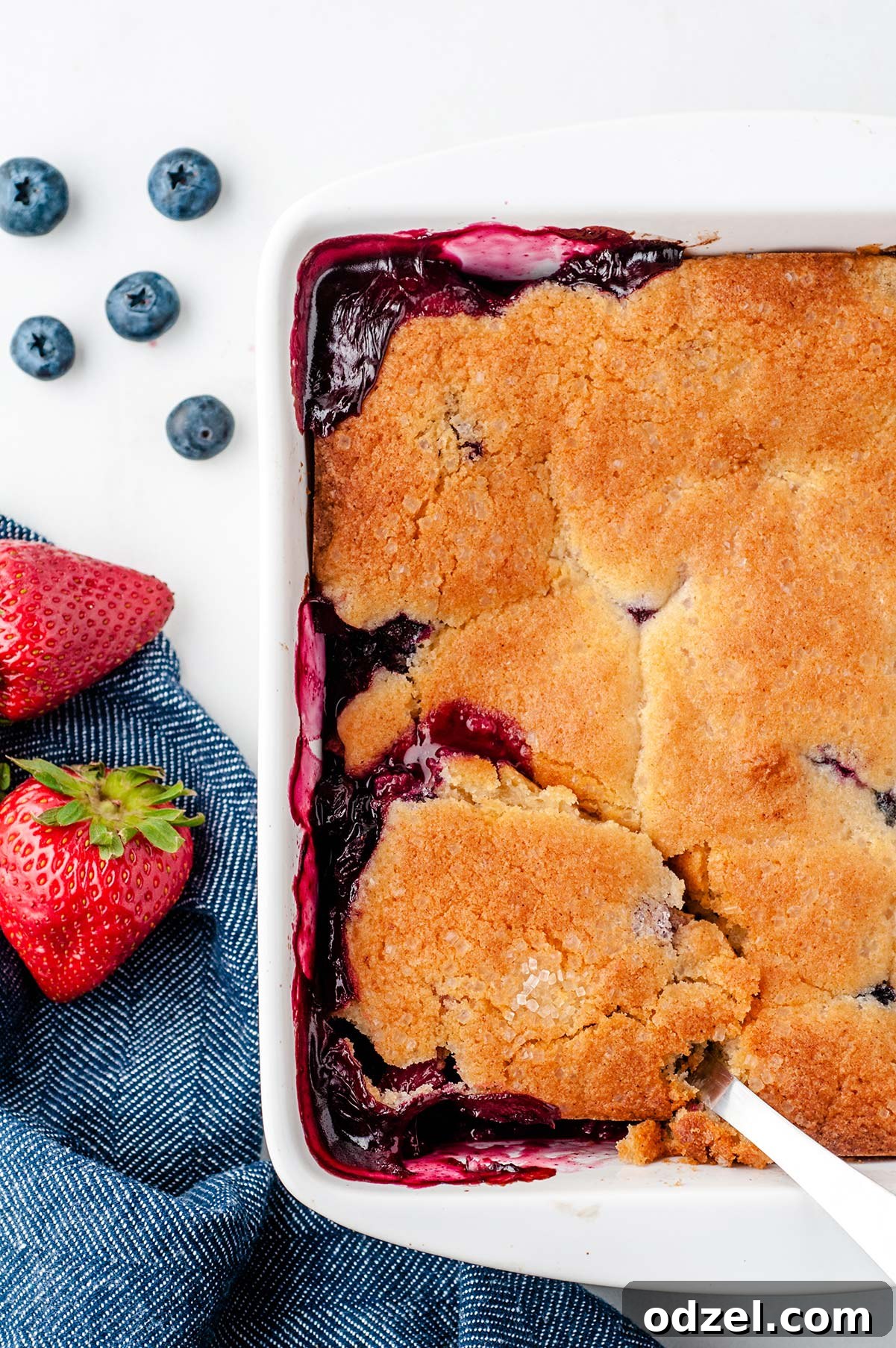 A close-up shot of half a white baking dish filled with a perfectly baked berry cobbler, showcasing its golden-brown topping and bubbling fruit. Fresh blueberries, strawberries, and a blue kitchen towel are arranged artfully on the side.