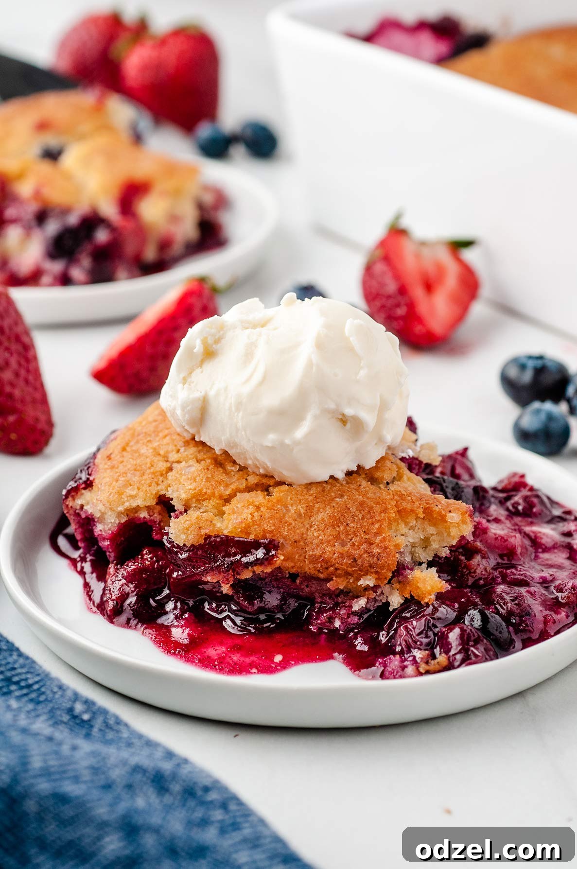 A close-up of a serving of berry cobbler on a white plate, topped with a melting scoop of vanilla ice cream. In the softly blurred background, a baking dish containing more cobbler and fresh berries is visible, enhancing the dessert's appeal.