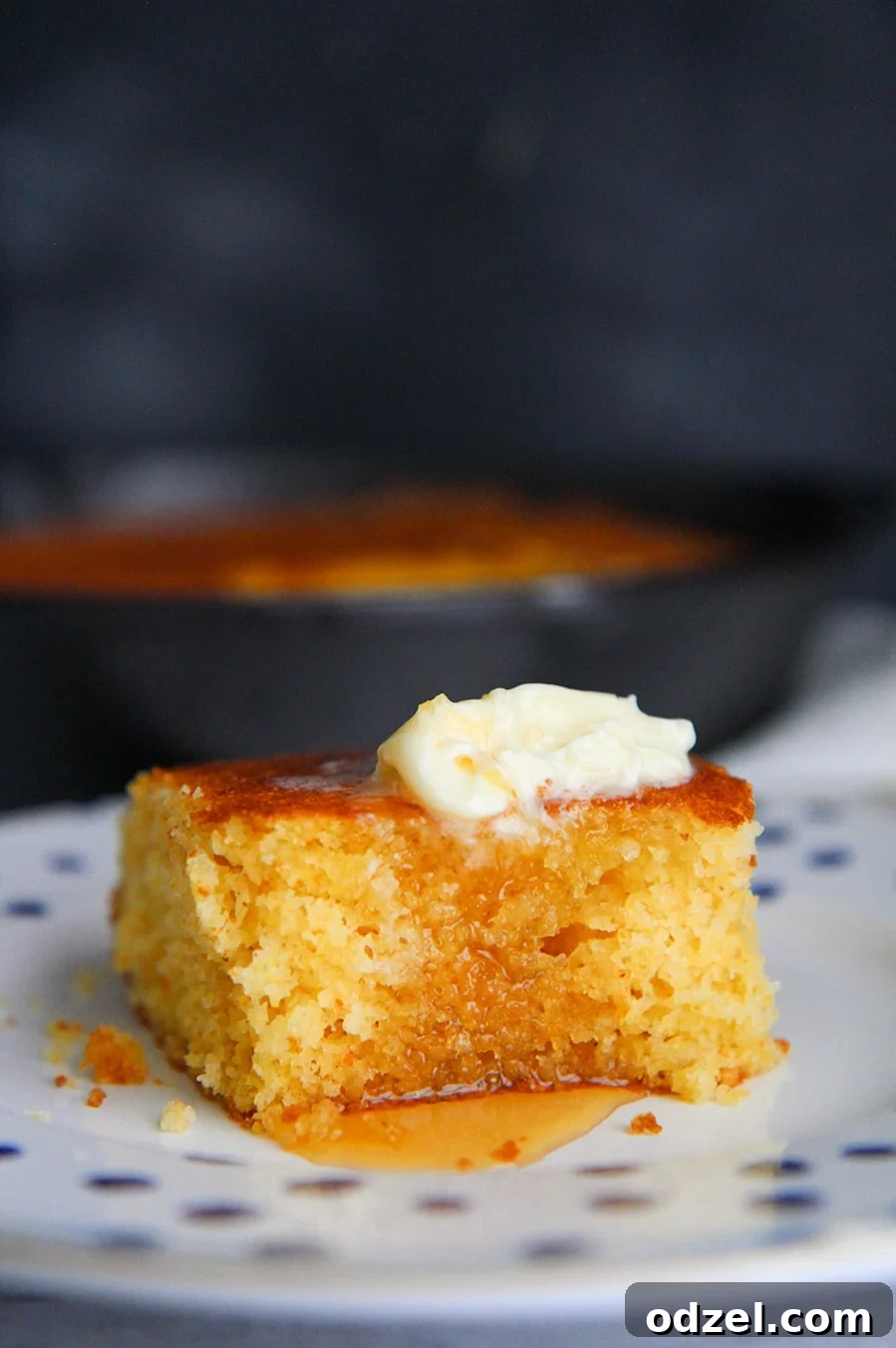 A golden-brown slice of buttermilk cornbread on a white plate, topped with melting butter and a drizzle of golden honey. The texture is invitingly moist and tender.