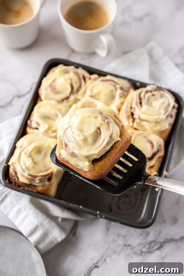 A spatula lifting a perfectly frosted cinnamon roll from a pan.