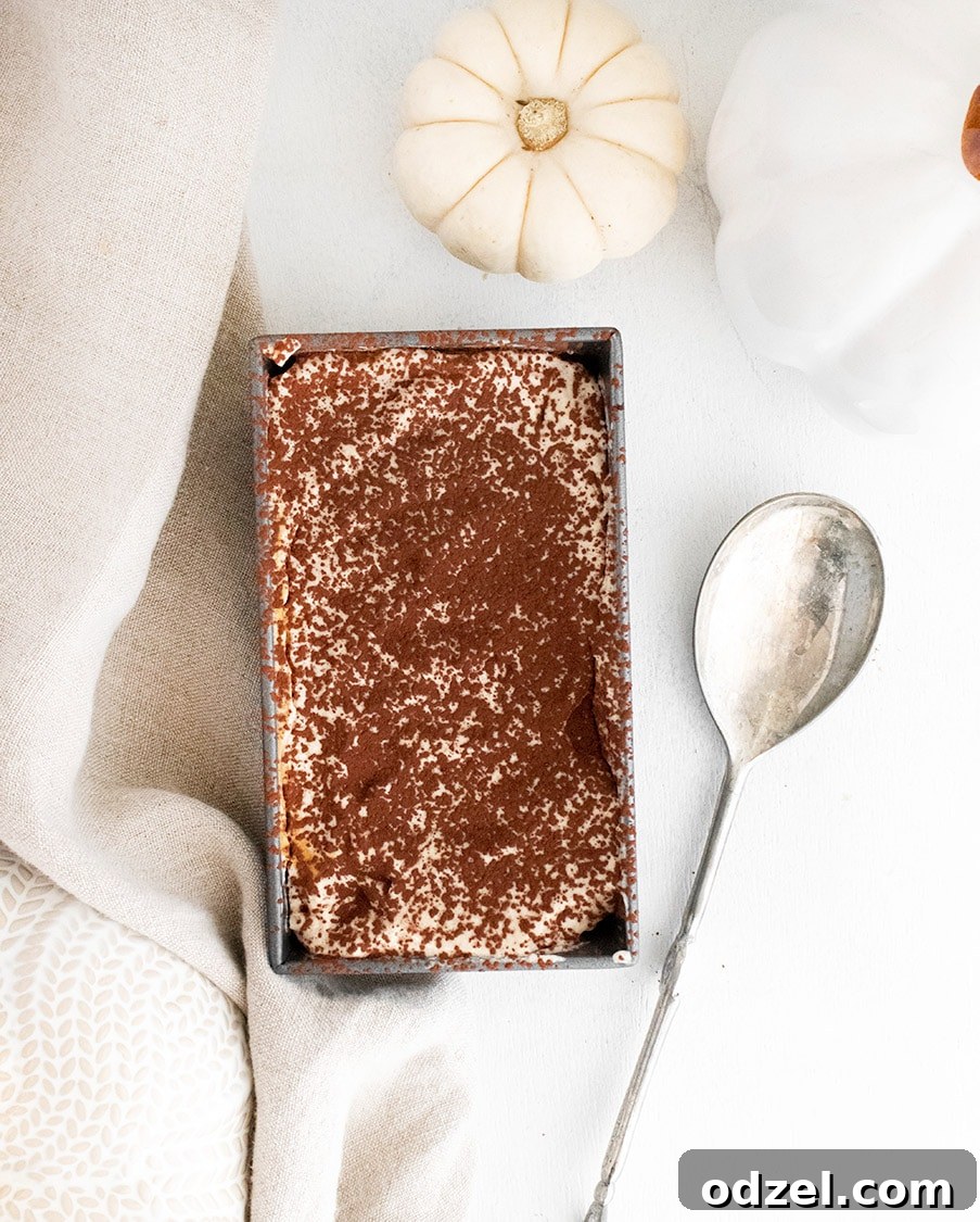 A dark-topped pumpkin tiramisu in a loaf pan, accompanied by a silver spoon, a light linen towel, and a small white pumpkin, ready for serving.