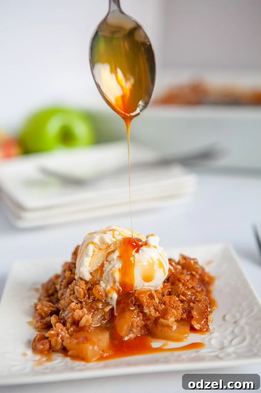 A spoon drizzling caramel sauce over warm apple crisp with melting vanilla ice cream on a white plate, ready to be enjoyed.