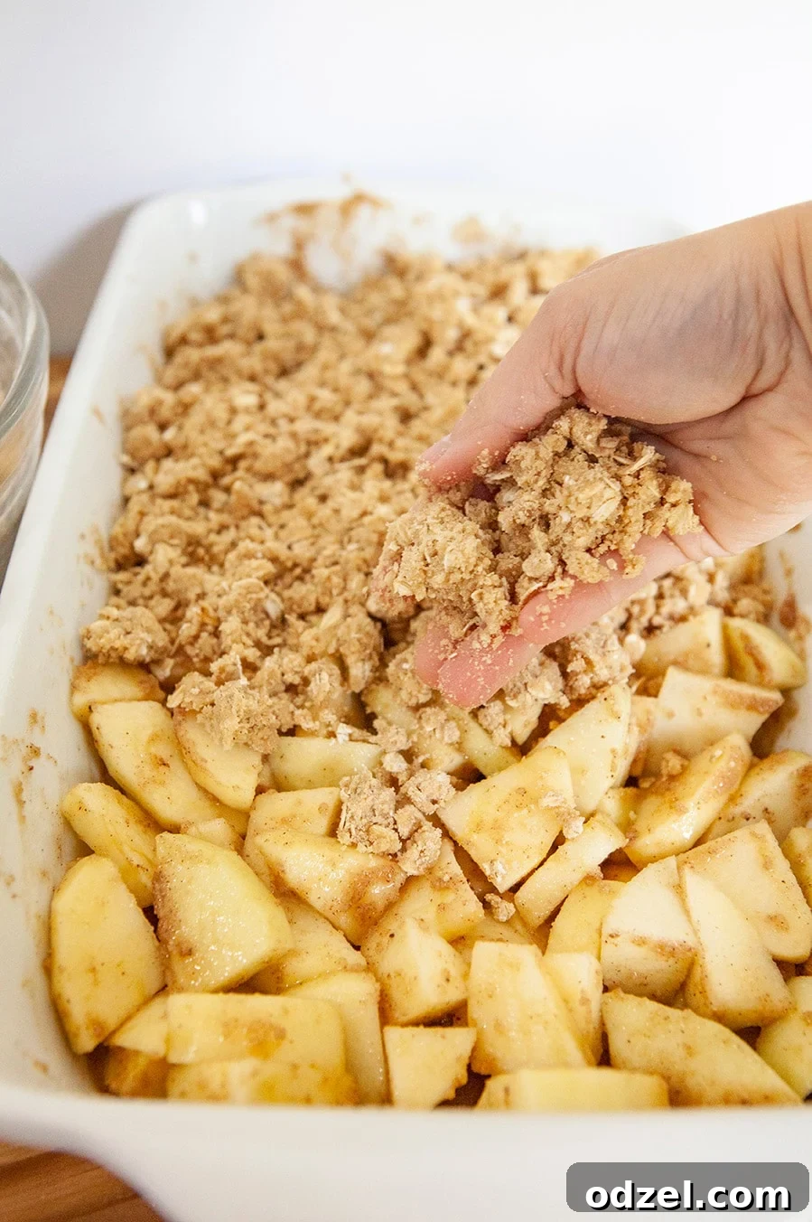 A hand pouring the crunchy oat topping mixture over thinly sliced apples in a white ceramic casserole dish before baking.