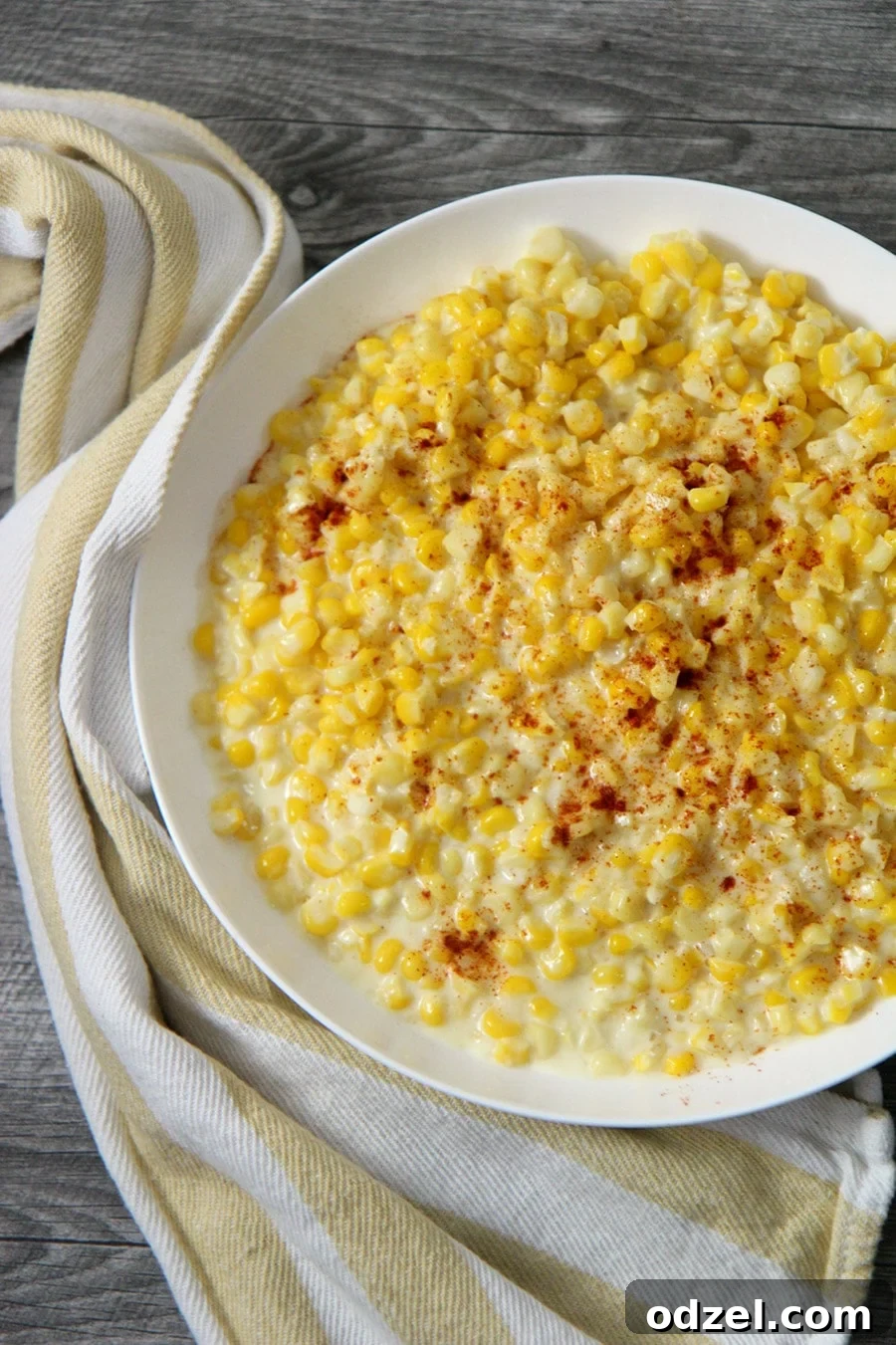 A white plate filled with creamy homemade creamed corn, garnished with fresh herbs, with a yellow and white kitchen towel elegantly draped beside it.