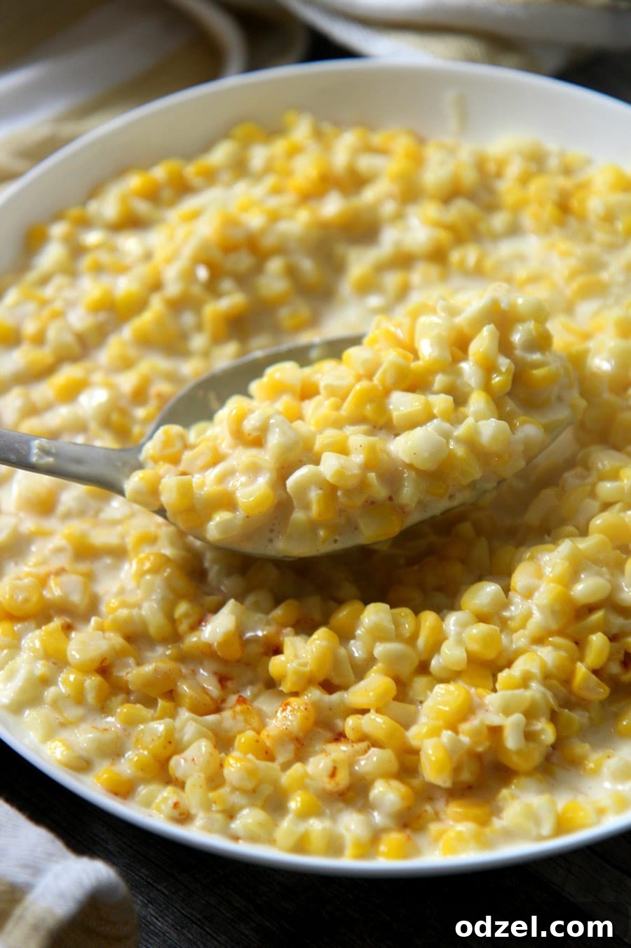 A serving spoon gracefully lifting a generous scoop of creamy, homemade creamed corn from a rustic white bowl, showcasing its rich texture and enticing appeal.