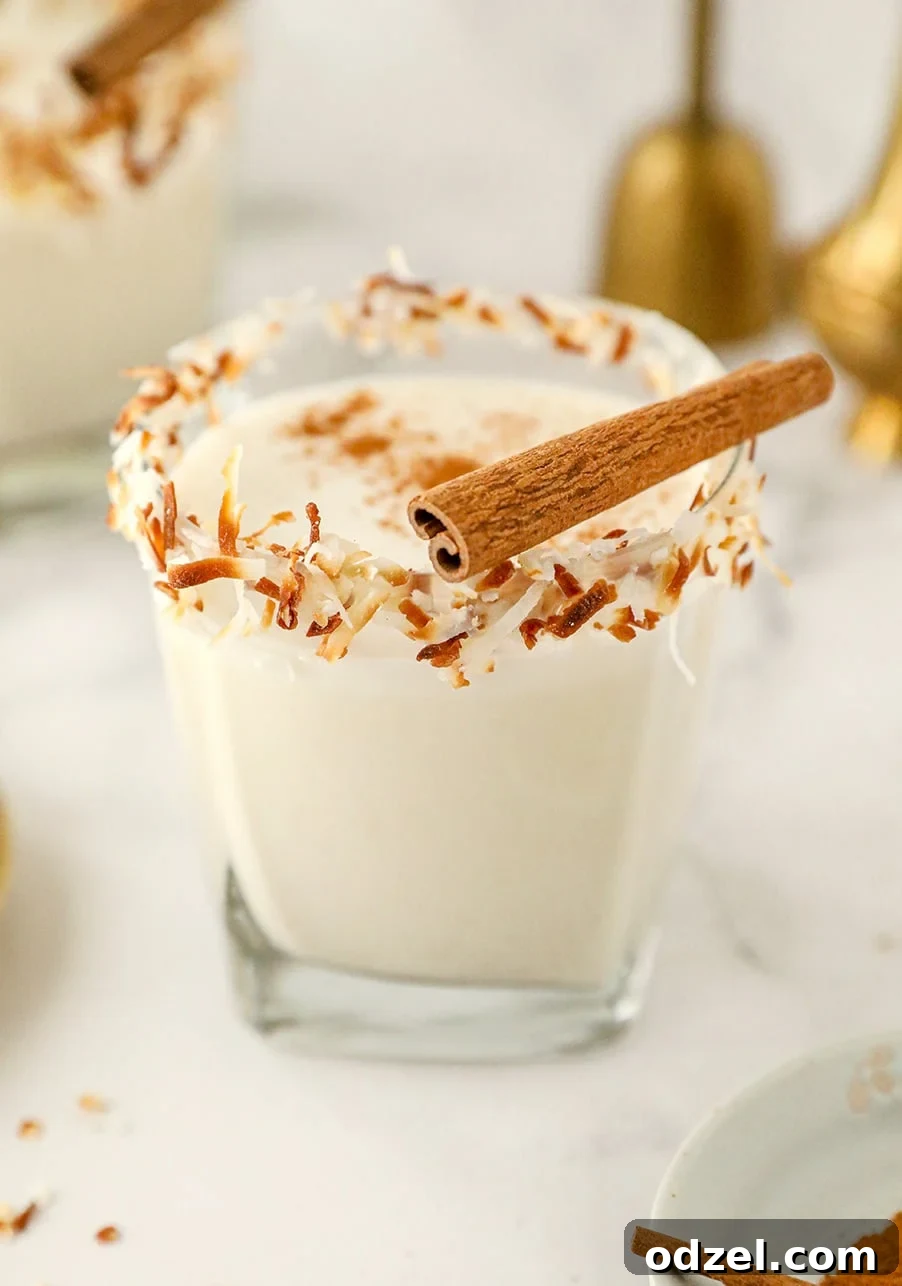 Coquito in a short glass with toasted coconut on the rim and a cinnamon stick on top.