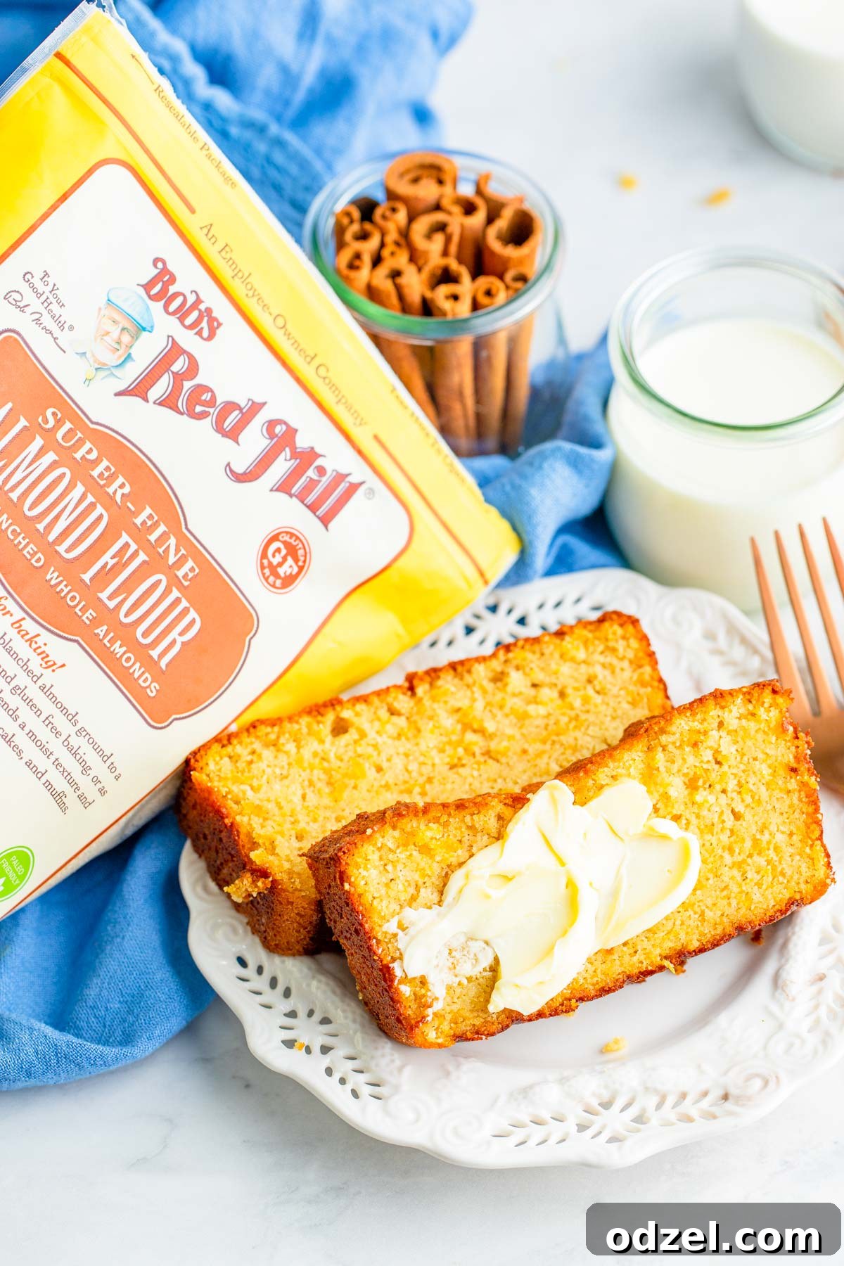Two slices of golden-brown sweet potato bread, one with a dollop of butter, alongside a fork, a glass of milk, and a package of Bob's Red Mill almond flour, highlighting a cozy breakfast scene.