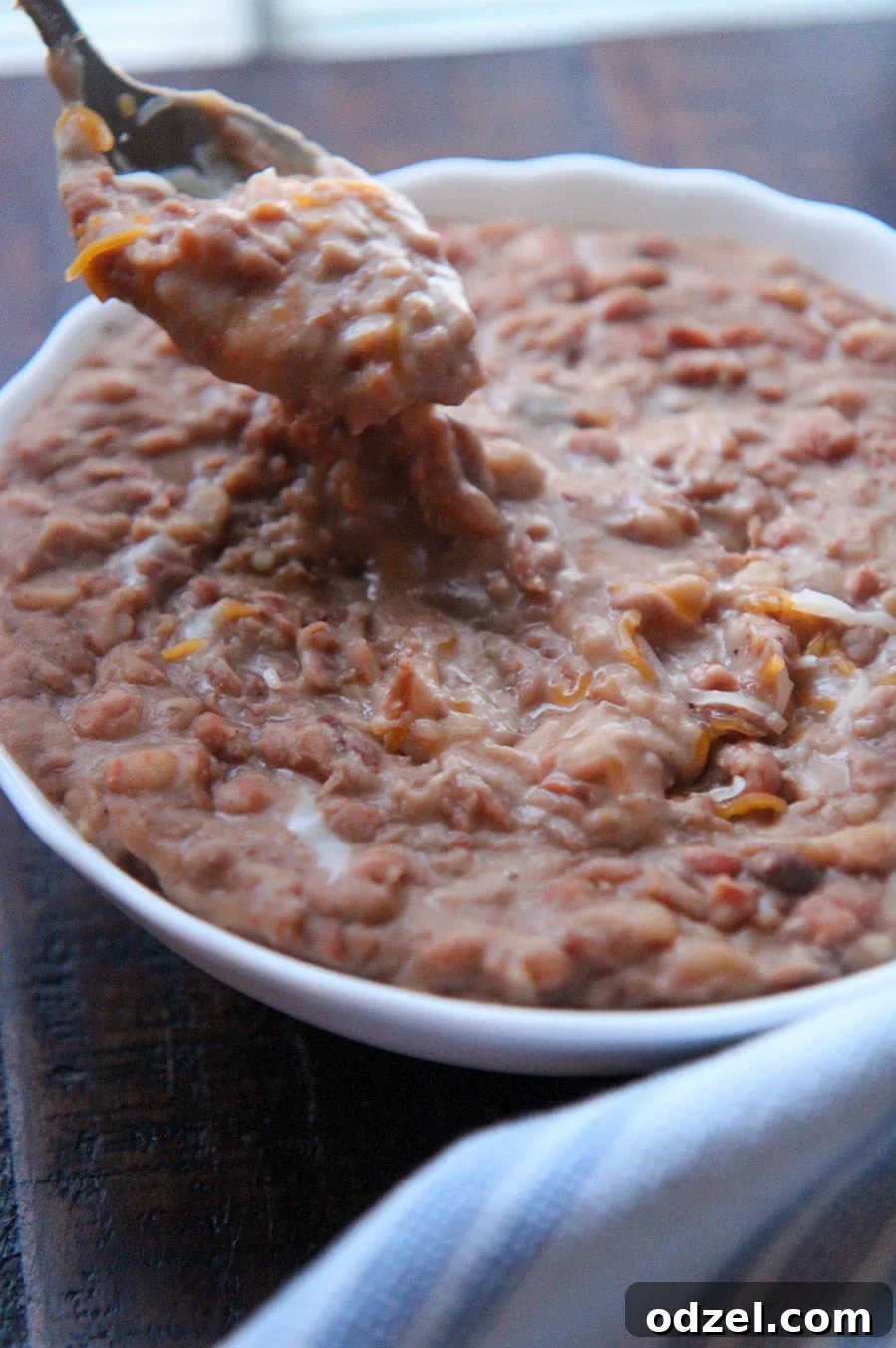 Creamy Instant Pot Refried Beans 5 Chunky refried pinto beans with glistening melted cheese, and a spoon dipped in the bowl, highlighting the texture.