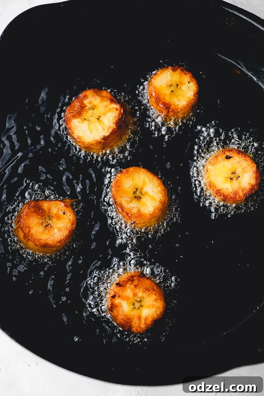 Several green plantain slices partially frying in hot oil within a cast iron skillet, turning golden brown.