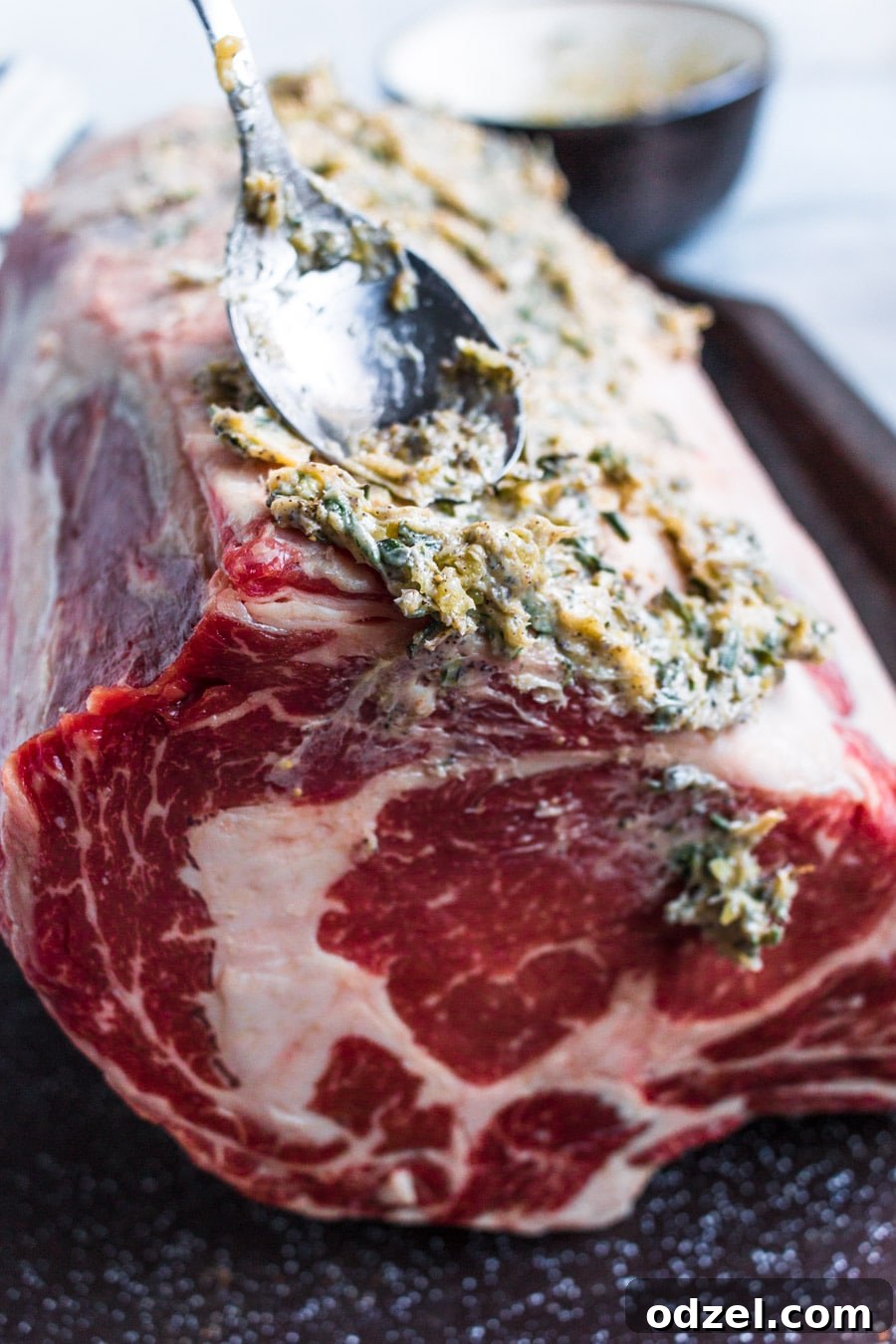 Juicy Prime Rib Masterpiece 3 Raw prime rib coated with a fresh herb butter rub, ready for roasting, with a spoon nearby.