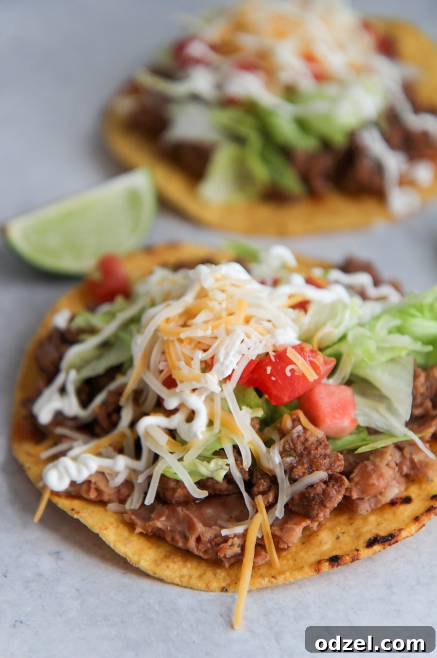 Two beef tostadas beautifully arranged with refried beans, crisp lettuce, diced tomatoes, shredded cheese, a dollop of sour cream, and fresh lime wedges on the side.