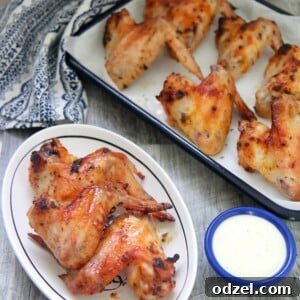 Oven roasted chicken wings on a small white plate with a side of blue cheese dip and more chicken wings in the background.