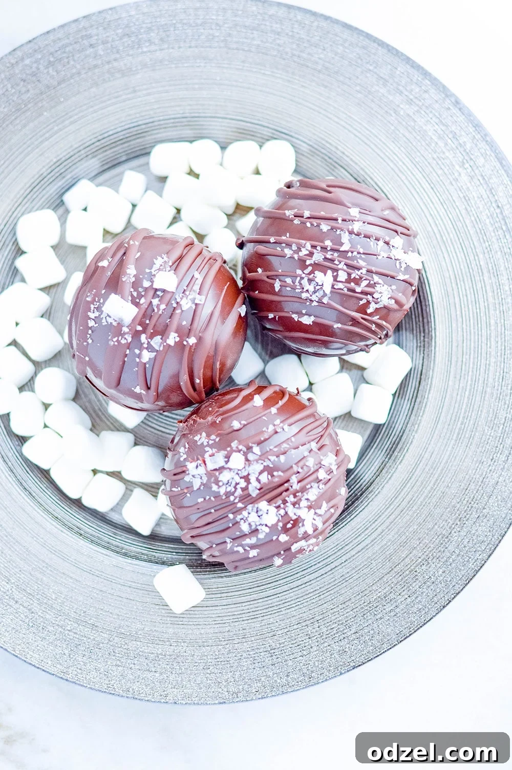 Three perfectly crafted salted caramel hot chocolate bombs resting on a small white plate, surrounded by scattered mini marshmallows, ready to be dropped into hot milk.