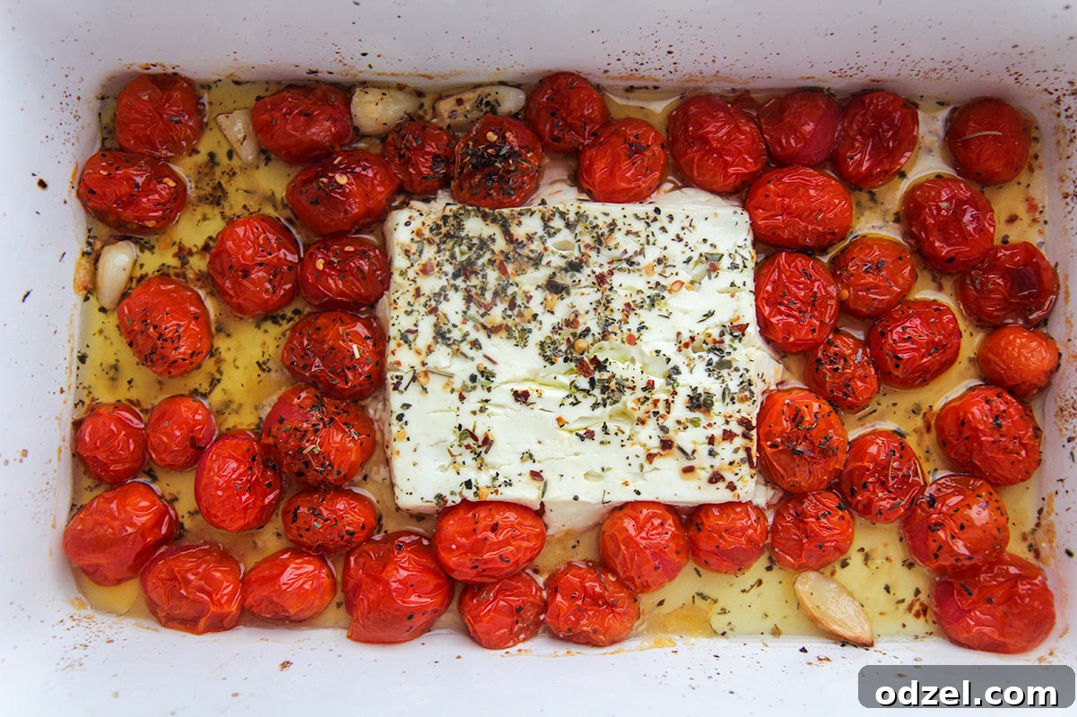 A block of feta cheese, olive oil, Italian seasoning, and cherry tomatoes arranged in a white baking dish, ready for baking.