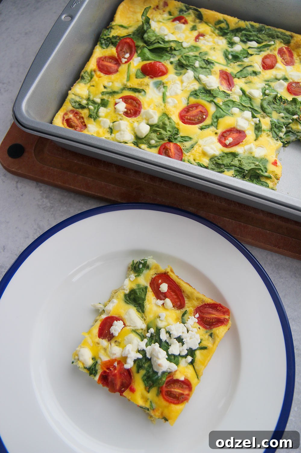 A beautifully sliced piece of golden-brown spinach and goat cheese frittata on a white and blue plate, with the full 9x13 casserole dish brimming with fresh ingredients in the soft-focused background, ready to be enjoyed.