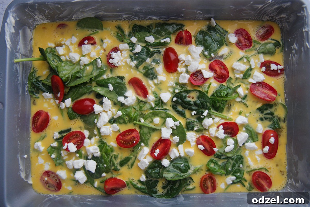 The raw, unbaked mixture of eggs, vibrant spinach, and creamy goat cheese filling a 9x13 buttered baking dish, topped with bright red cherry tomatoes, ready for the oven to transform it into a delicious frittata.