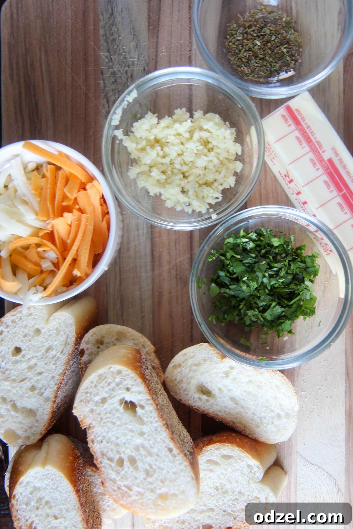 cheese, minced garlic, and fresh herbs in small bowls, alongside a baguette and softened butter on a wooden board.