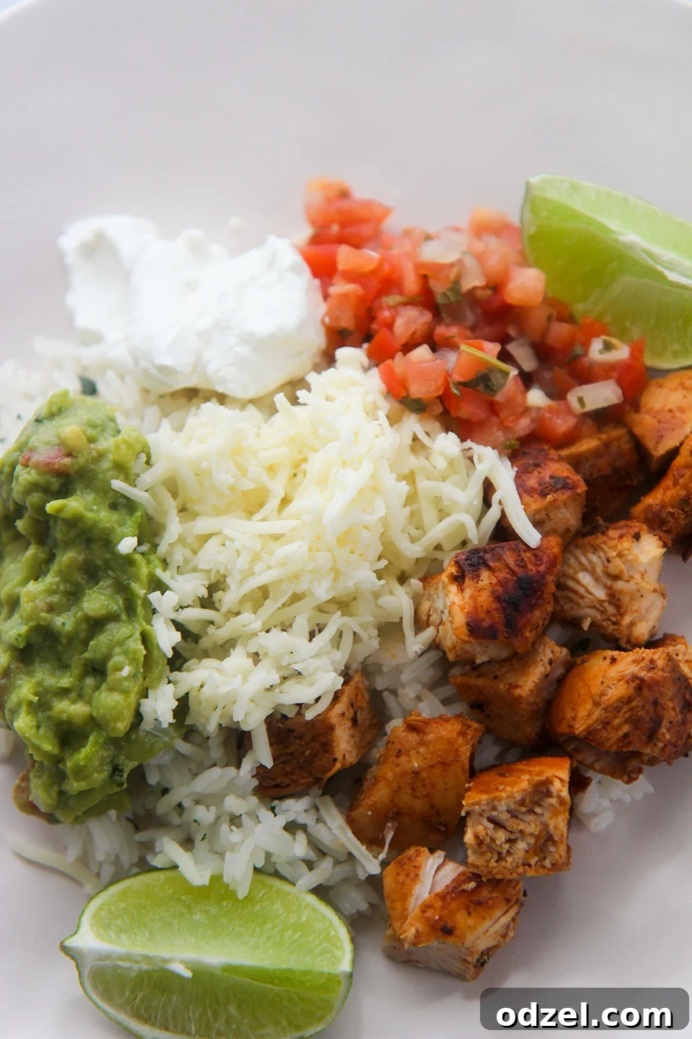 A vibrant and colorful Chipotle-style bowl featuring guacamole, shredded cheese, fresh pico de gallo, tender chipotle chicken, sour cream, and bright lime wedges on a white plate, showcasing a complete meal.