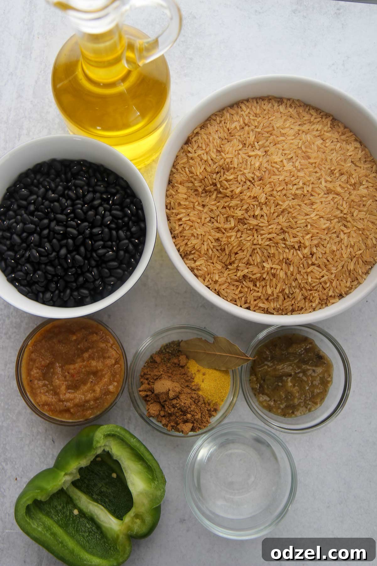 A vibrant display of key ingredients for making Cuban Congri. Dried black beans are ready for cooking, alongside brown rice, small bowls filled with aromatic spices like cumin and oregano, a bottle of vinegar, bay leaves, homemade sofrito, and recaito. A bottle of olive oil and a fresh green bell pepper complete the setup for this flavorful Cuban dish.