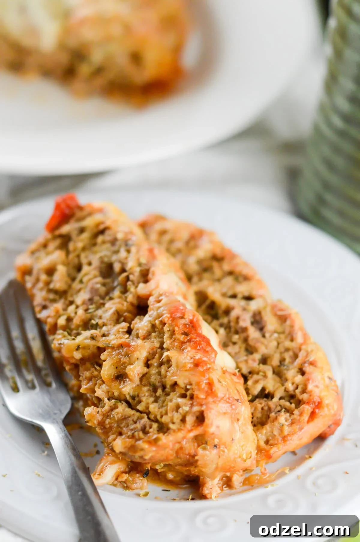 Two generous slices of Meatloaf Parmesan served on a pristine white plate, garnished with fresh herbs, with a fork resting beside it. The cheese is melted and slightly browned, and the texture of the meatloaf is clearly visible, promising a tender bite.