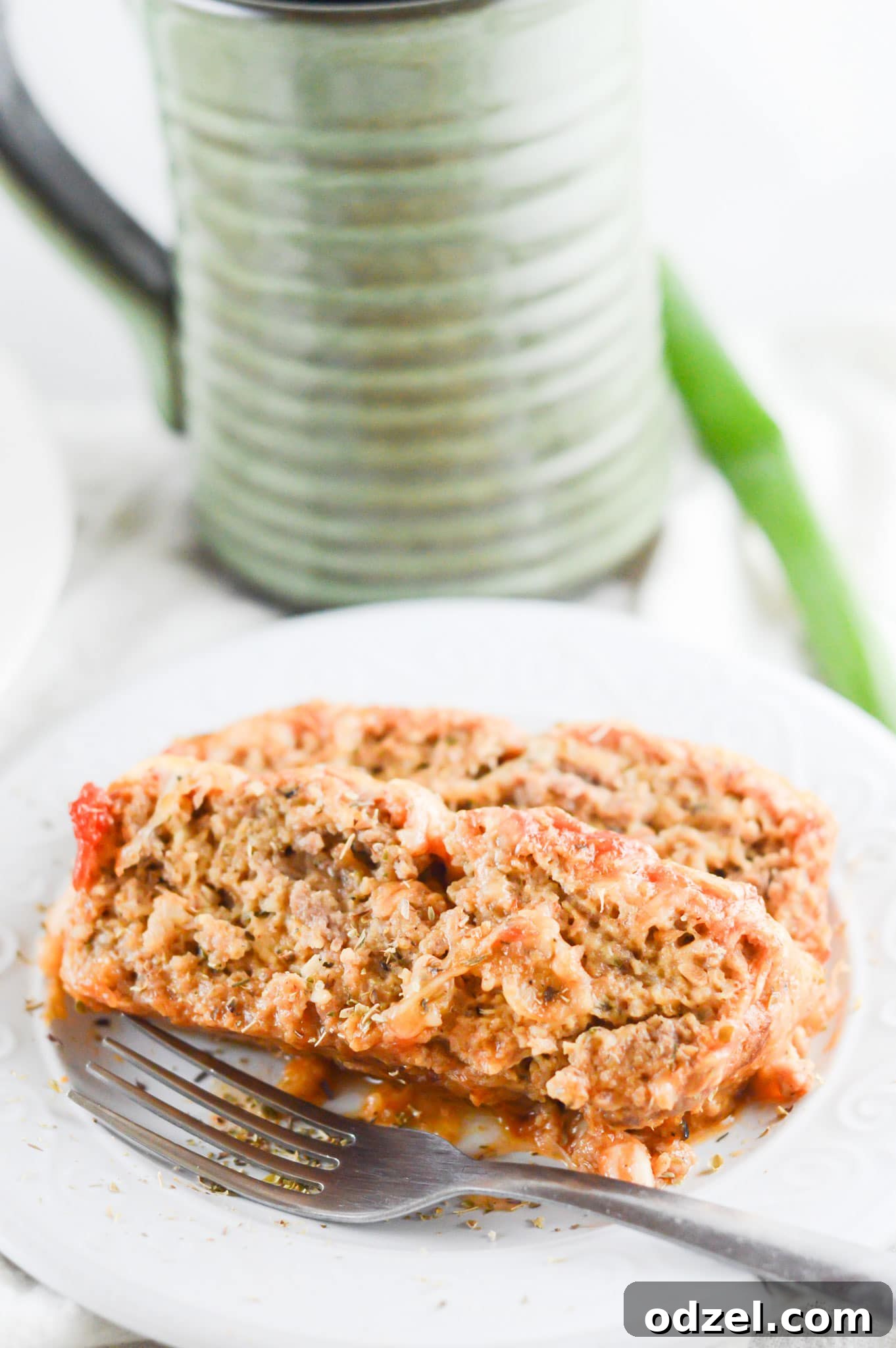 Two perfectly sliced portions of Meatloaf Parmesan, glistening with melted cheese and marinara, presented on a white plate. A fork rests alongside, and a beverage in a glass cup is visible in the soft-focus background, suggesting a complete and satisfying meal.