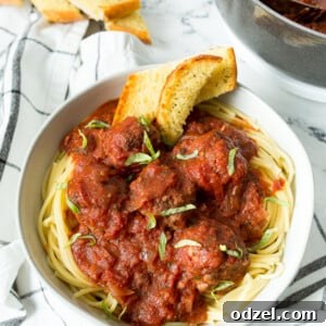 A white bowl brimming with spaghetti and meatballs, accompanied by golden garlic bread, set on a blue and white patterned towel. A pot of meatballs simmers in the background.