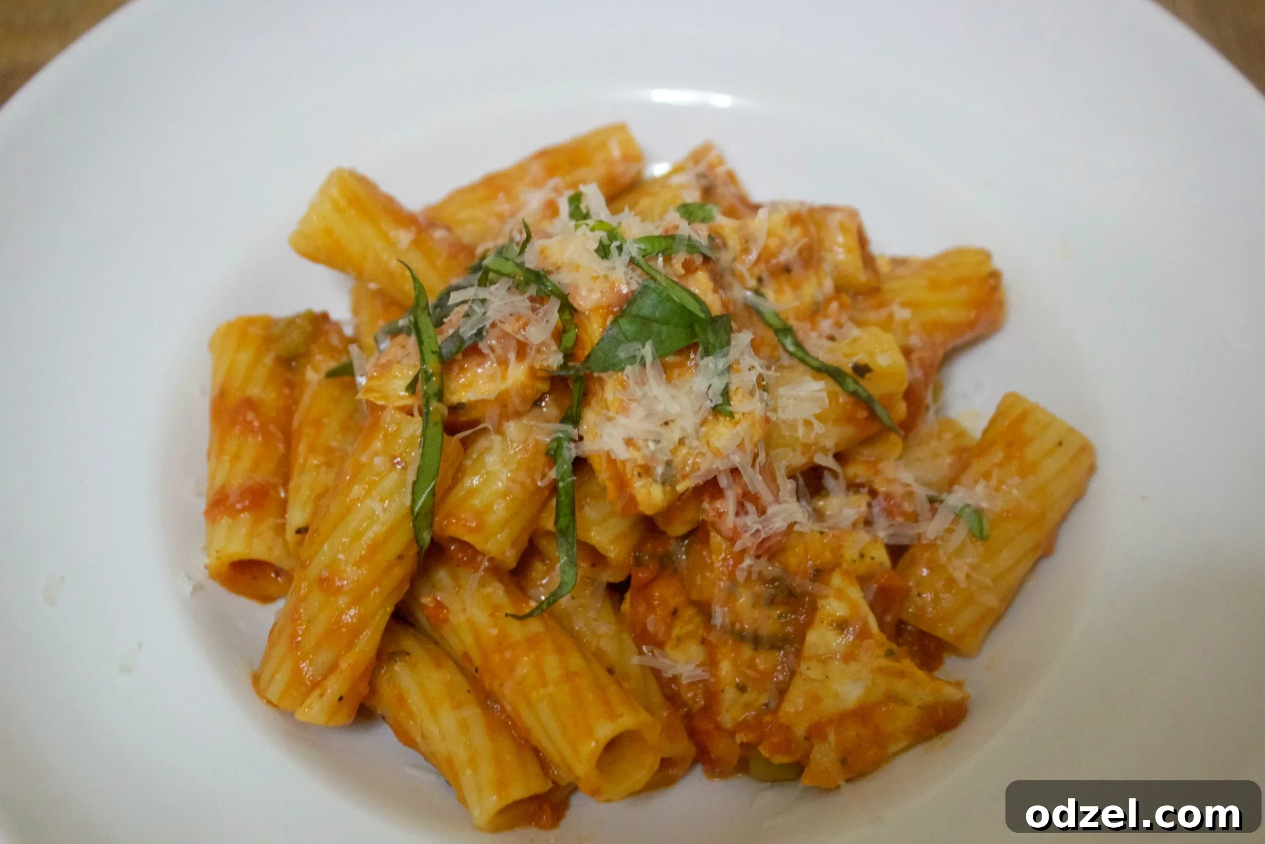 Fiery Chicken Rigatoni 2 A vibrant bowl of spicy chicken rigatoni, adorned with fresh parmesan cheese and basil leaves. The pasta is coated in a rich, reddish sauce with succulent chicken pieces and green peas.